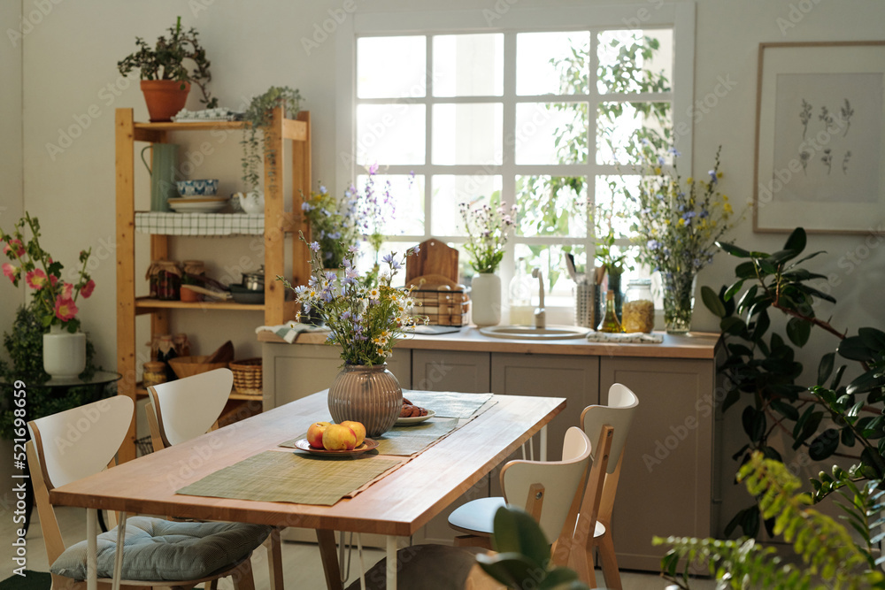 Cozy kitchen with wooden table in the center surrounded by sink with kitchenware and wildflowers by window and green domestic plants