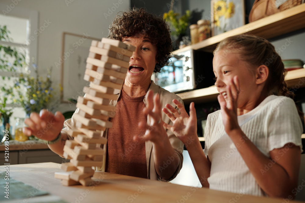 Young woman and her youthful daughter trying to hold on fallen tower built up of wooden blocks while sitting by table during game Young woman and her youthful daughter trying to hold on fallen tower built up of wooden blocks while sitting by table during game