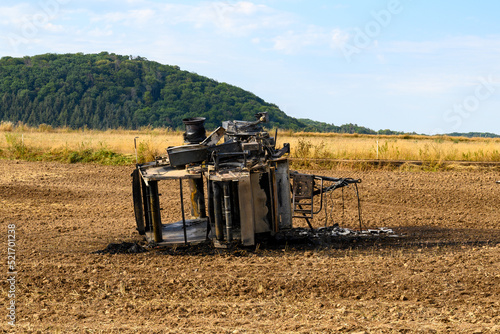 Top view of a burnt out hay baler lying in a field against a green forest