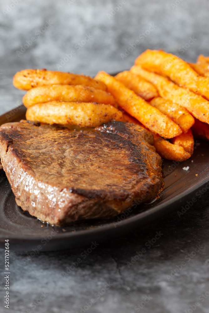 Rump steak dinner, with sweet potato fries and onion rings, on a black oval plate.  On a dark stone background
