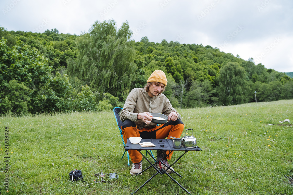 A tourist guy sits at a table in nature dining on a hike, trekking in the mountains, morning scrambled eggs in a frying pan, wild conditions, camping in the forest. A tourist guy sits at a table in nature dining on a hike, trekking in the mountains, morning scrambled eggs in a frying pan, wild conditions, camping in the forest.