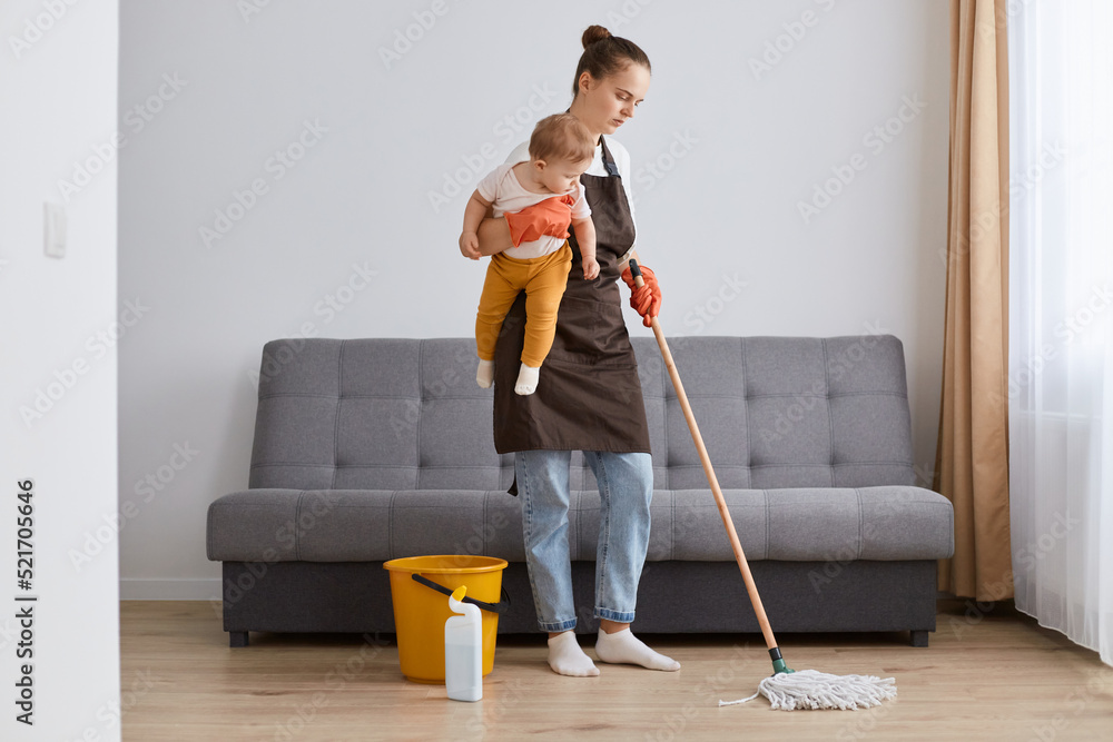Fototapeta premium Horizontal shot of tired exhausted woman wearing brown apron with mop and little toddler daughter in hands, washing floor, tiding up apartment, cleaning in living room.
