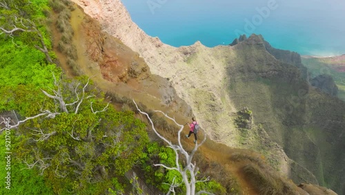 Camera follows young woman hiking with backpack and trekking poles on top of jungle mountain summit at sunset or sunrise. Outdoor adventure trip in wilderness Napali coast park on Kauai Hawaii island