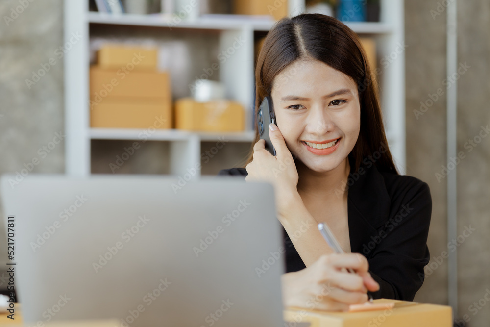 Asian woman is talking on the phone with a customer to confirm an order, she owns an online store, she packs and ships through a private transport company. Online selling and online shopping concepts. Asian woman is talking on the phone with a customer to confirm an order, she owns an online store, she packs and ships through a private transport company. Online selling and online shopping concepts.