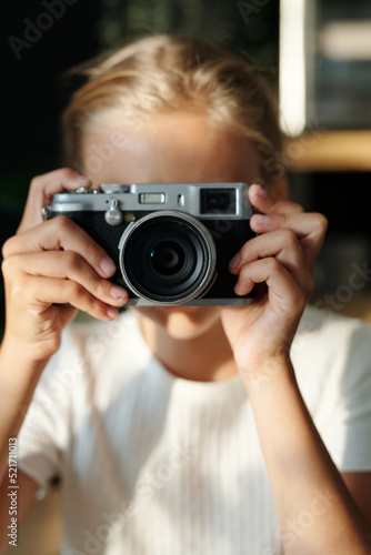 Hands of youthful blond girl holding retro photocamera by her face while taking shots of still life or people standing in front of her