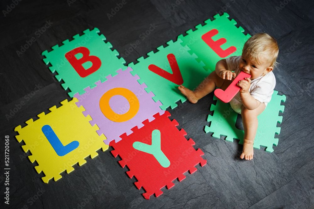 Naklejka premium bird view of colorful kids puzzle mat playground in nursery with letters like Baby Love written on them lying on the floor while one year old blond baby in white body is playing 