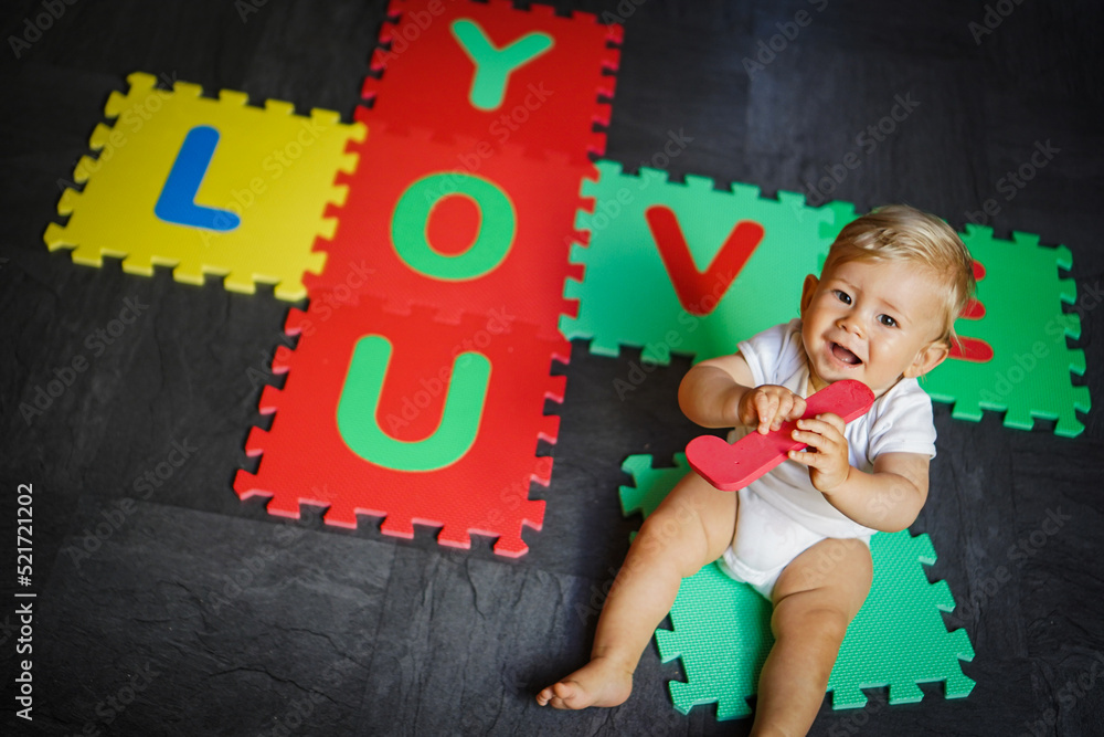 Naklejka premium bird view of colorful kids puzzle mat playground in nursery with letters like Baby Love written on them lying on the floor while one year old blond baby in white body is playing 