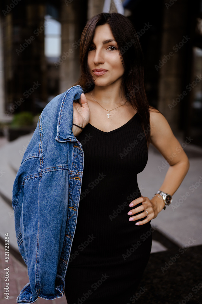 Fashionable young brunette woman in black dress and long jeans jacket on the city street. Fashion . Stylish silver women's wrist watch on a model's hand .