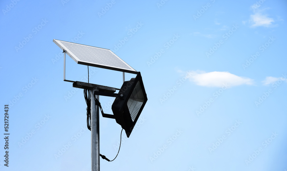 Mini photovoltaic or solar cell panel and floodlight led installed on metal pole against blue sky to store and save the energy from the sun to use the light at night around the house or the park. Mini photovoltaic or solar cell panel and floodlight led installed on metal pole against blue sky to store and save the energy from the sun to use the light at night around the house or the park.