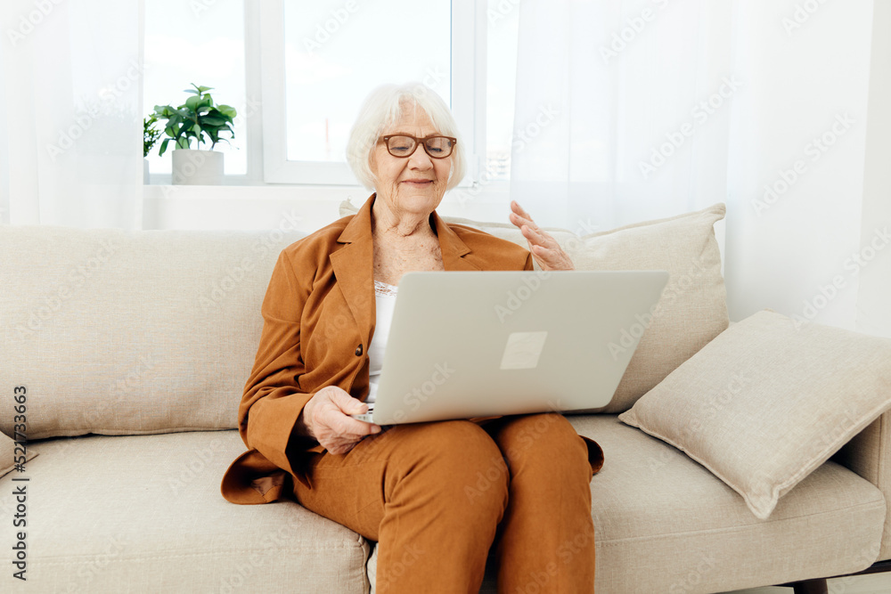 a joyful, happy old lady in a brown suit is sitting on a beige sofa having fun communicating with loved ones via video link holding a laptop on her lap a joyful, happy old lady in a brown suit is sitting on a beige sofa having fun communicating with loved ones via video link holding a laptop on her lap