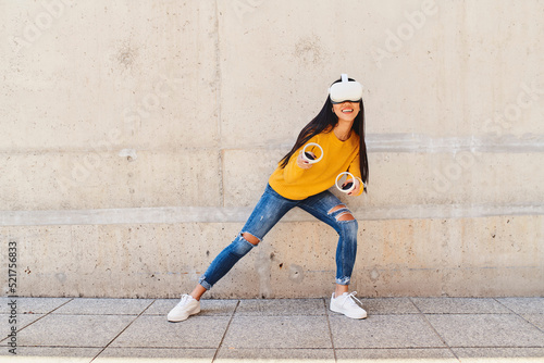 Young woman sneaking while playing with vr goggles standing against concrete wall