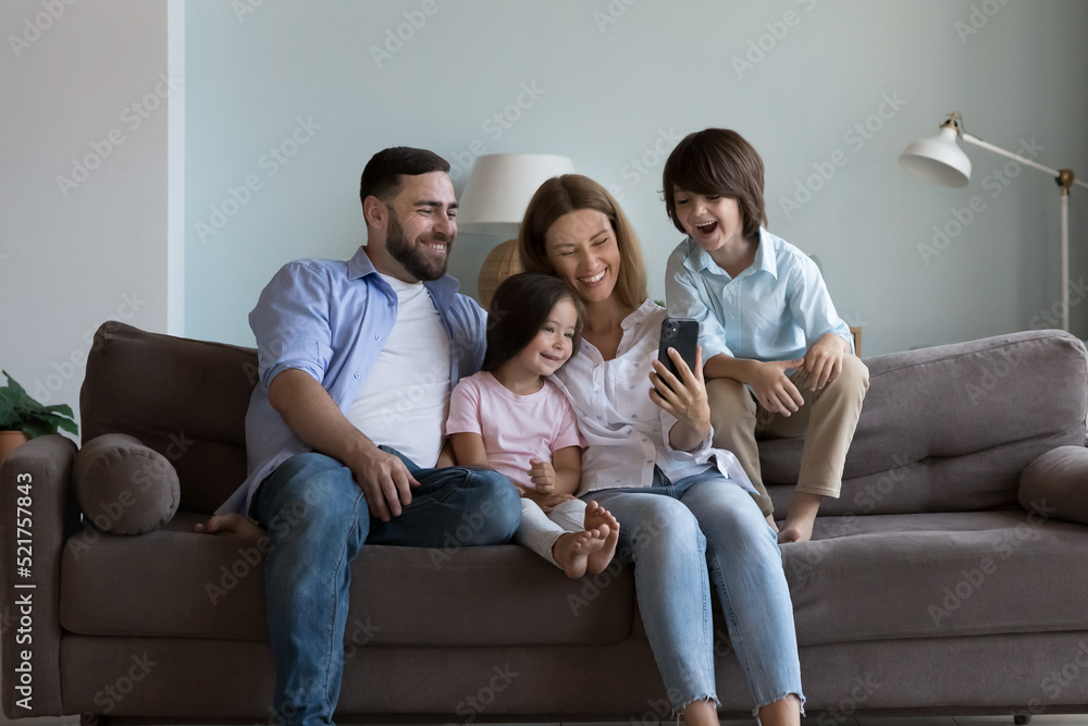 Cheerful children and young parents have fun watch videos on internet sit on sofa use smartphone feel happy enjoy new mobile application, laughing spend carefree leisure together at home. Tech concept Cheerful children and young parents have fun watch videos on internet sit on sofa use smartphone feel happy enjoy new mobile application, laughing spend carefree leisure together at home. Tech concept