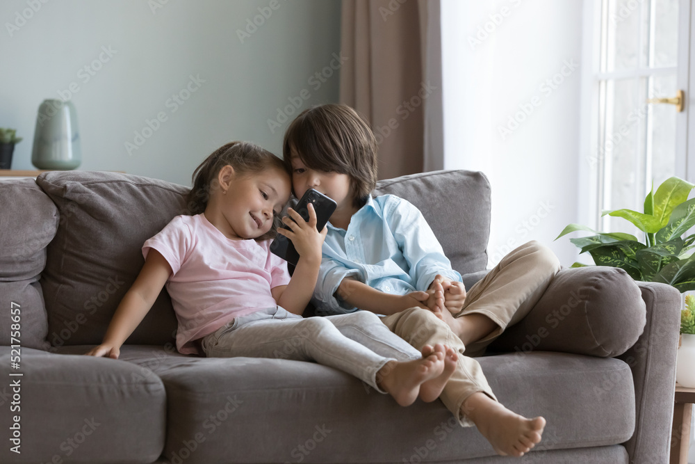 Cute preschoolers kids use smartphone at home. Little pretty boy and girl spend leisure hold modern cellphone sit on sofa in living room. Watch vlog on-line content, play games. Gadget overuse concept