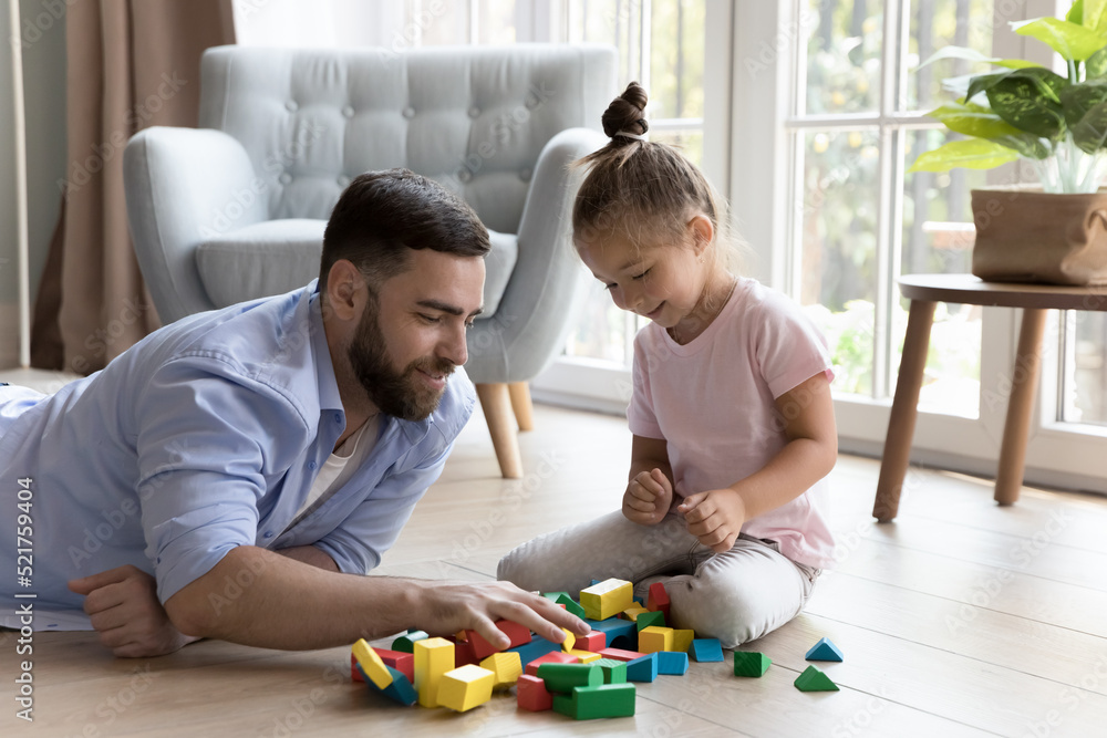 Young daddy and cute little 4s daughter build together with colored wooden block settled down on warm floor with underfloor heat, in cozy living room. Kids development, family leisure, pastime concept Young daddy and cute little 4s daughter build together with colored wooden block settled down on warm floor with underfloor heat, in cozy living room. Kids development, family leisure, pastime concept
