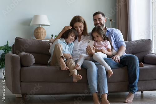 Family with little children sit on couch look at smart phone screen enjoy video conference, virtual meeting with grandparents, spend leisure using mobile app, young generation and tech usage concept