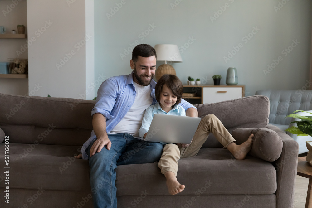 Dad and son sit on sofa enjoy weekend use laptop, rest at home spend leisure on internet, buying goods, make order on-line, choose cartoons or family movies on digital streaming services. Tech concept