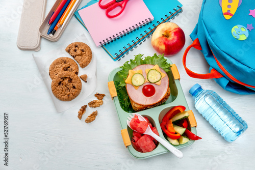 School lunch box for kids with food in the form of funny faces. School lunch box with sandwich, vegetables, water and stationery on table. Healthy eating habits concept. Back to school concept photo