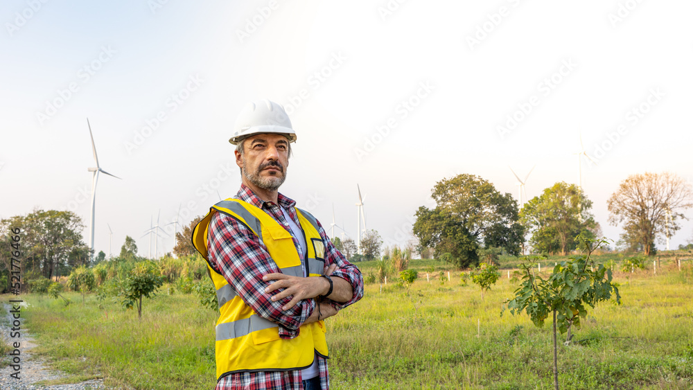Aืn Engineer standing by the wind turbine in the renewable power farm. He is wearing a white hard hat and yellow transparent vest. Aืn Engineer standing by the wind turbine in the renewable power farm. He is wearing a white hard hat and yellow transparent vest.
