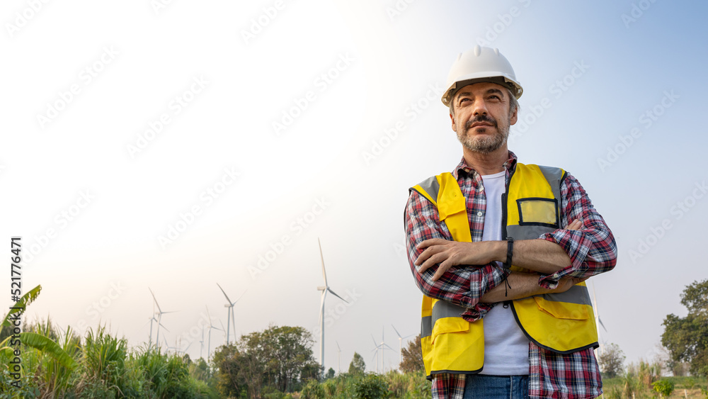 Aืn Engineer standing by the wind turbine in the renewable power farm. He is wearing a white hard hat and yellow transparent vest. Aืn Engineer standing by the wind turbine in the renewable power farm. He is wearing a white hard hat and yellow transparent vest.