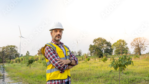 A   n Engineer standing by the wind turbine in the renewable power farm. He is wearing a white hard hat and yellow transparent vest.