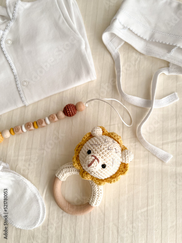 A baby's white suit, a hat for a newborn, scratches, socks, a rattle with a lion and a chain of wooden beads for teeth are lying on the bed. vertical photo