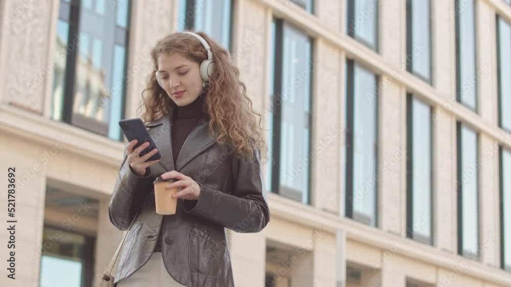 Low angle medium slowmo of attractive young smiling woman in headphones scrolling on smartphone standing outdoors against tall beige building with takeaway coffee cup in hand Low angle medium slowmo of attractive young smiling woman in headphones scrolling on smartphone standing outdoors against tall beige building with takeaway coffee cup in hand