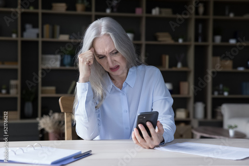 Frustrated annoyed elderly retired woman holding wrong working mobile phone, getting error message, spam problem, bad news, low signal, connection trouble, feeling negative emotions photo