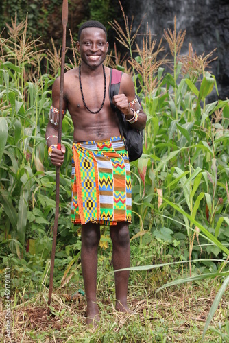 Education in Africa,farming,agriculture: A barefoot young African Masai Moran warrior student stands holding a backpack and spear 