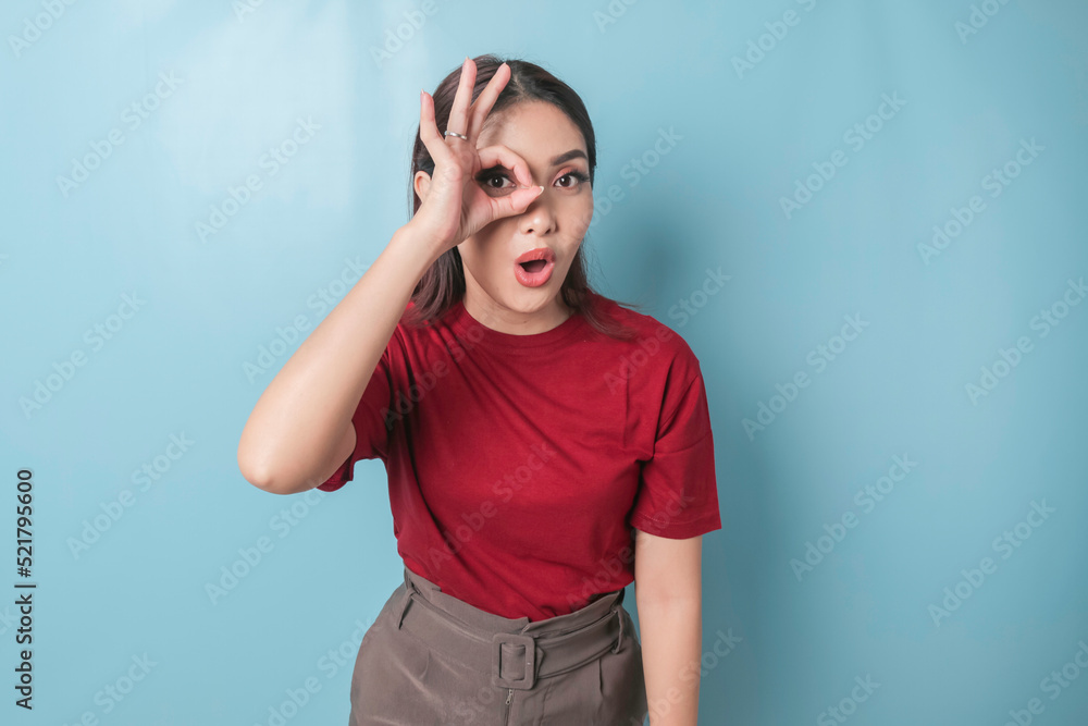 Excited Asian woman wearing a red t-shirt giving an OK hand gesture isolated by a blue background