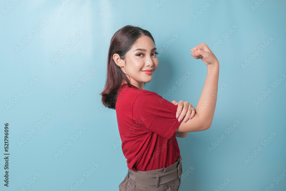 Fototapeta premium Excited Asian woman wearing a red t-shirt showing strong gesture by lifting her arms and muscles smiling proudly