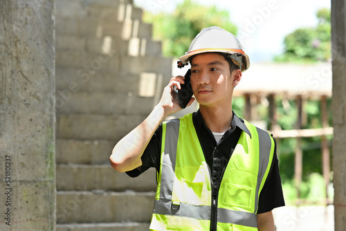 Young Asian engineer, architect or foreman wearing safety helmet and reflective clothing talking on smartphone while looking forward.