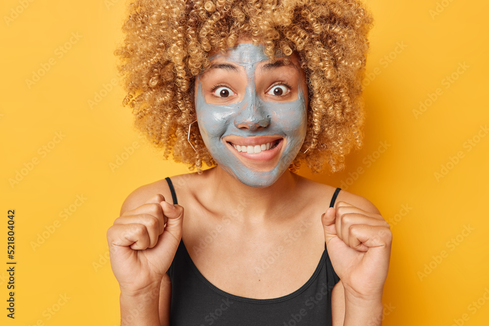 Positive surprised woman with curly hair clenches fists bites lips looks excited at camera applies beauty mud mask wears black t shirt isolated over vivid yellow background. Wellness and cosmetology