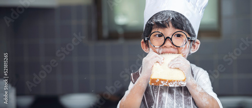 Cute asian boy wearing chef hat apron eating bread in kitchen at home
