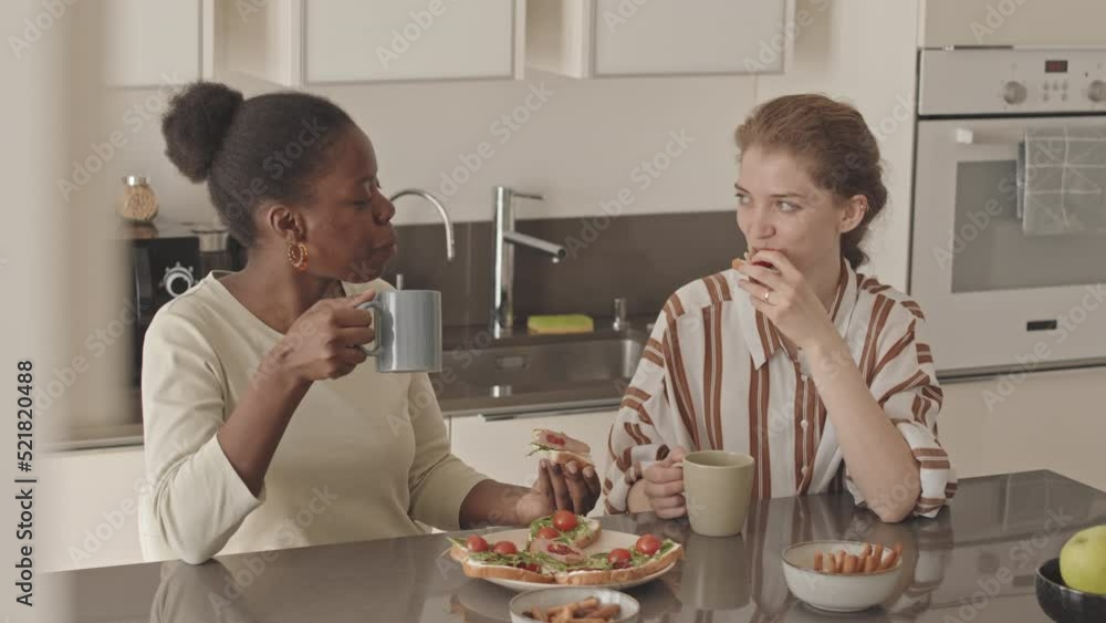 Medium slowmo of delighted young diverse lesbian couple chatting while eating homemade sandwiches at table in modern minimalist style kitchen Stock ビデオ | Adobe Stock