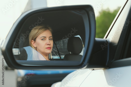 Reflection of an attractive woman in a car rearview mirror. A blonde woman with bright make-up is driving a car. Portrait of a serious woman. © Galina