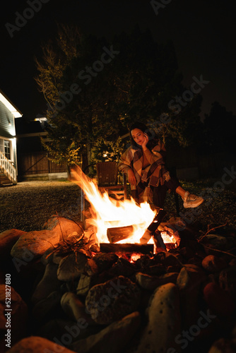 A woman covered with a plaid sits by the fire. Backyard of a wooden house at night.