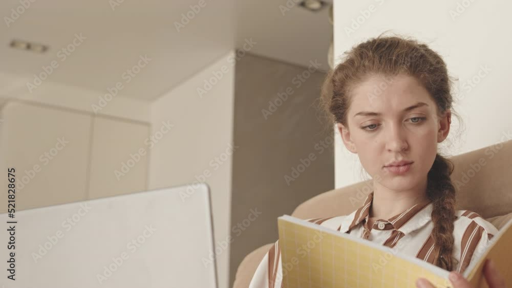 Waist up slowmo of concentrated young Caucasian businesswoman working on laptop on sofa at home while house maid cleaning apartment in background Waist up slowmo of concentrated young Caucasian businesswoman working on laptop on sofa at home while house maid cleaning apartment in background