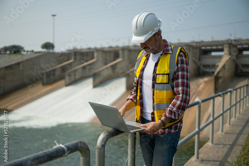 Portrait of engineer wearing yellow vest and white helmet with laptop computer Working day on a water dam with a hydroelectric power plant. Renewable energy systems, Sustainable energy concept photo