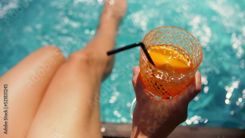 Woman soaking her feet in swimming pool and holding orange cocktail.Young woman drink cocktail near swimming pool.Sexy woman sit near swimming pool relaxing vacation.