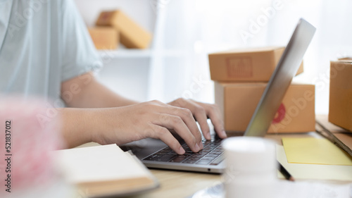 Young man uses a laptop to chat with customers who come to order product, Freelance work at home, Packaging on background, Sell online, Small business owner, Online shopping SME entrepreneur.