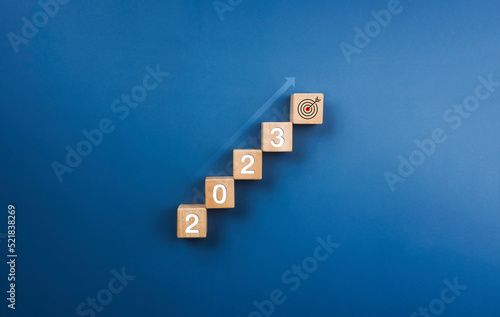 2023 year numbers on on wooden blocks stack, chart steps with shining rise up arrow to the target icon on blue background, business growth process to success, and economic improvement concept of 2023.