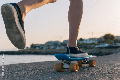Side close-up image of a young guy's foot riding a penny longboard by the sea or promenade at sunset. Alternative concept of sport among middle-aged people.