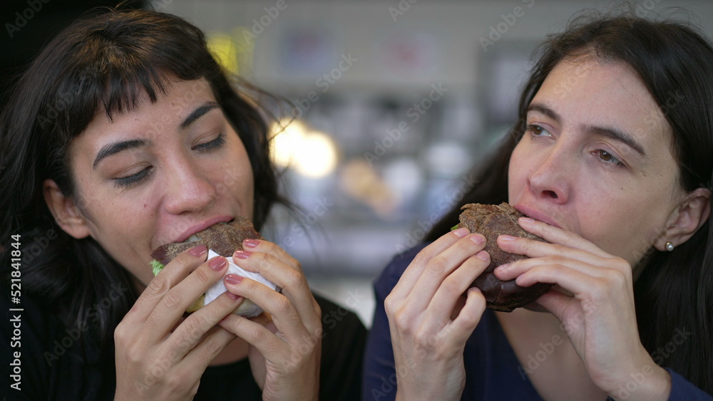 Two happy female friends eating burgers at the same time. Young women taking a bite of cheeseburgers at restaurant Two happy female friends eating burgers at the same time. Young women taking a bite of cheeseburgers at restaurant