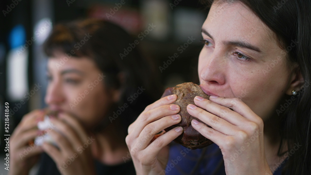 Two people eating burgers. Young women taking a bite of cheeseburgers. Female friends eating fast food lunch Two people eating burgers. Young women taking a bite of cheeseburgers. Female friends eating fast food lunch