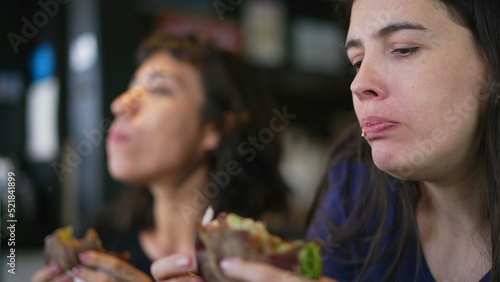 Two people eating burgers. Young women taking a bite of cheeseburgers. Female friends eating fast food lunch