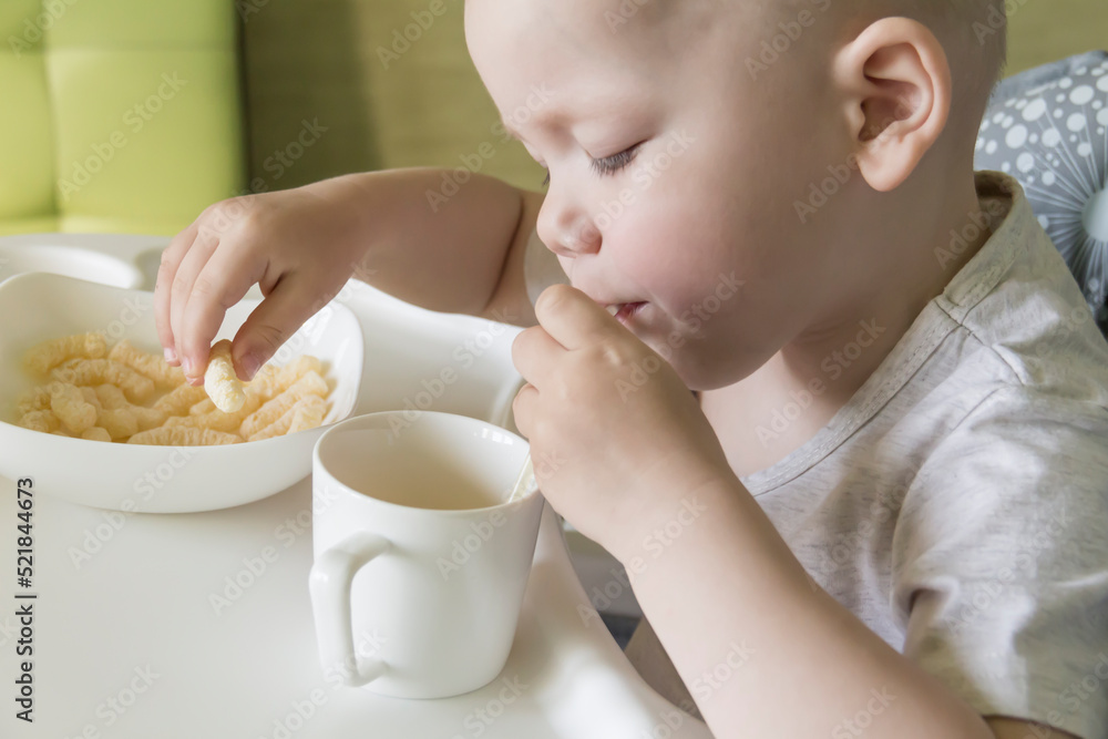 A small child with a short haircut crunches appetitively with corn sticks and drinks juice from a straw. A small child with a short haircut crunches appetitively with corn sticks and drinks juice from a straw.