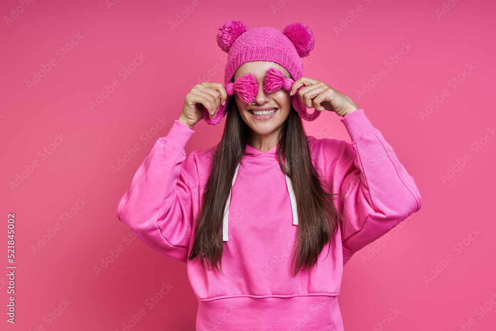 Joyful young woman in hooded shirt playing with her funky hat against pink background Joyful young woman in hooded shirt playing with her funky hat against pink background