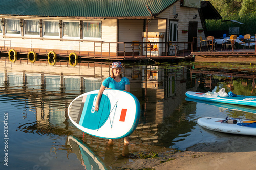 A young girl in a full cap and a blue T-shirt is holding a paddle board under her arm.