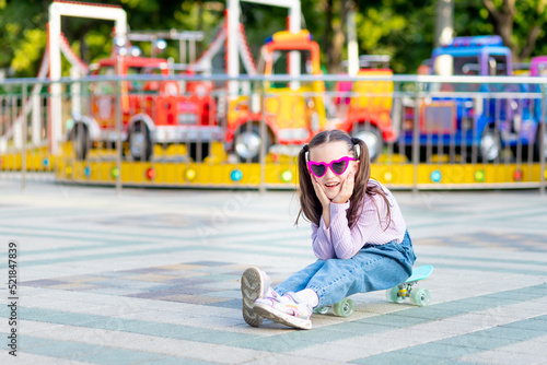a child girl in an amusement park rides a skateboard in the summer and smiles with happiness near the carousels in sunglasses, the concept of summer holidays and school holidays