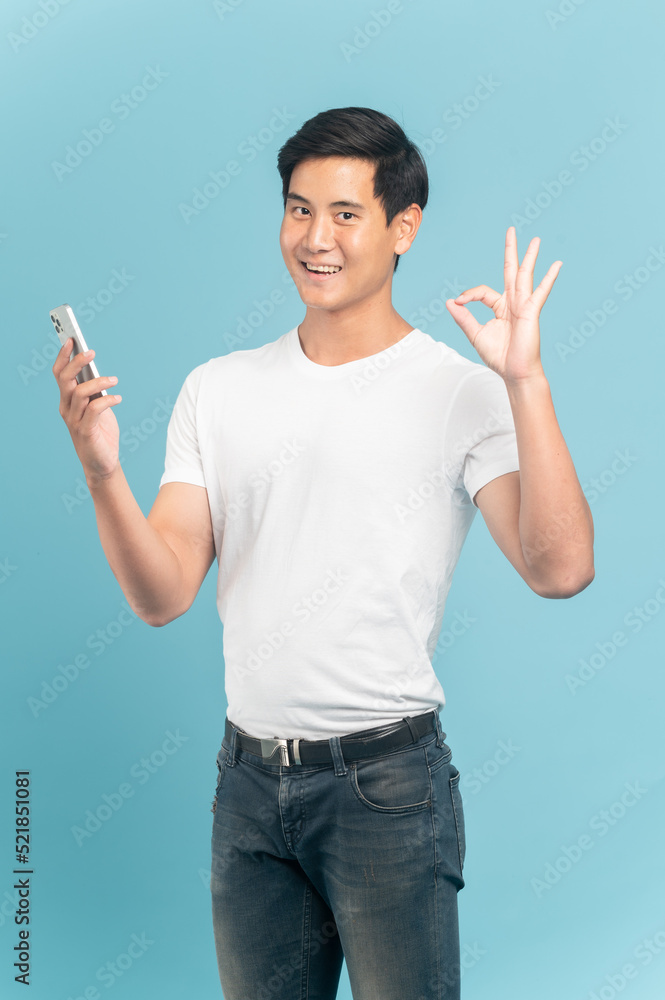 Nice asian guy in white t-shirt and jeans holding smartphone and make ok sign isolated on blue background Nice asian guy in white t-shirt and jeans holding smartphone and make ok sign isolated on blue background